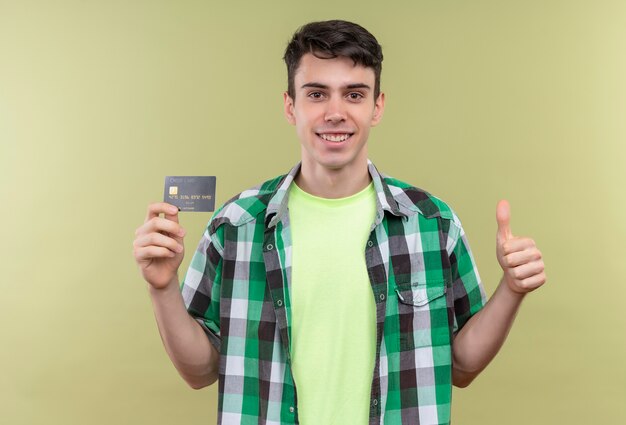 Glimlachende Kaukasische jonge kerel die groen overhemd draagt dat creditcard zijn duim op geïsoleerde groene achtergrond houdt