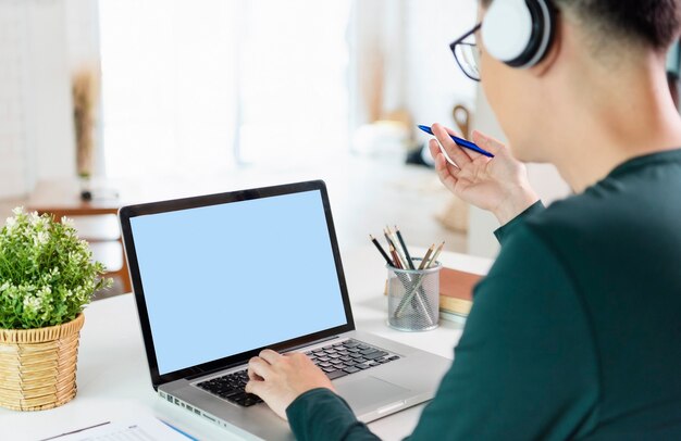 Glimlachende knappe Aziatische zakenman draagt een koptelefoon die op afstand werkt vanuit huis. Hij is webinar videoconferentie, Laptop leeg scherm.