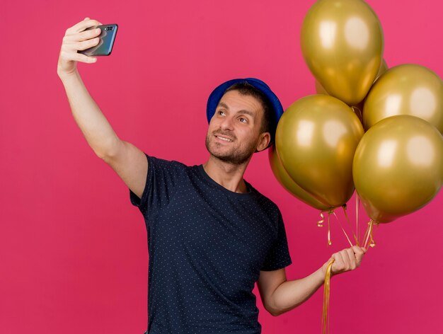 Glimlachende knappe blanke man met blauwe feestmuts houdt helium ballonnen nemen selfie kijken naar telefoon geïsoleerd op roze achtergrond met kopie ruimte