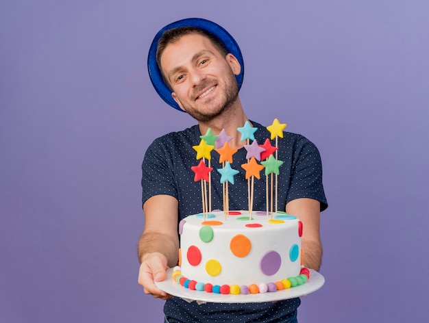 Glimlachende knappe blanke man met blauwe hoed houdt verjaardagstaart kijken camera geïsoleerd op paarse achtergrond met kopie ruimte
