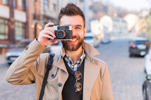 Glimlachende knappe jonge mens op stadsstraat die een beeld van uitstekende camera nemen