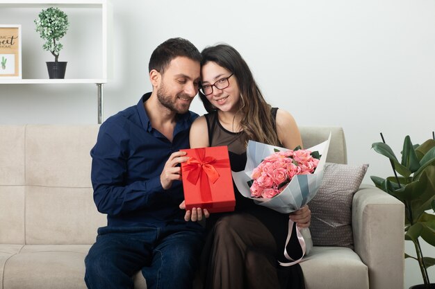 Glimlachende knappe man die een geschenkdoos geeft aan een tevreden mooie jonge vrouw in een bril met een boeket bloemen op de bank in de woonkamer