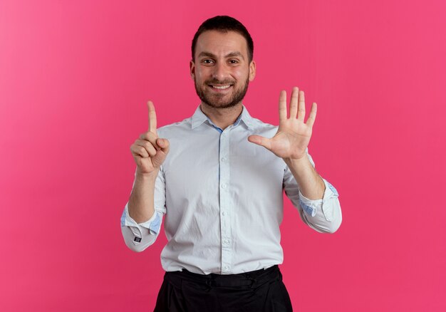 Glimlachende knappe man gebaren zes met handen geïsoleerd op roze muur