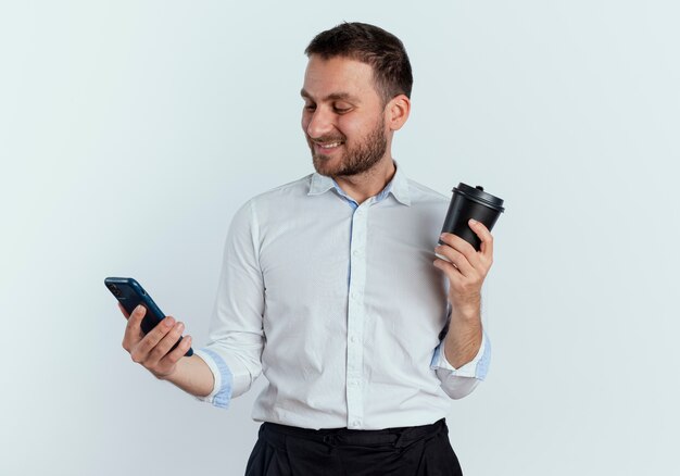 Glimlachende knappe man houdt koffiekopje en kijkt naar telefoon geïsoleerd op een witte muur