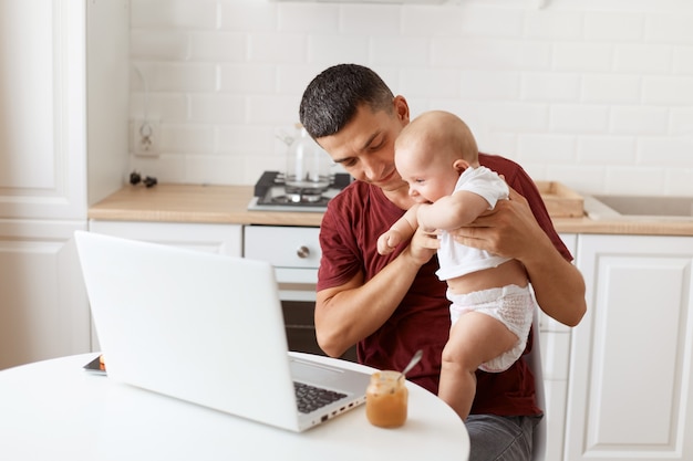 Glimlachende knappe man met donker haar met een bordeauxrood casual t-shirt, aan het werk op een laptop terwijl hij aan het babysitten en spelen is met zijn dochter, poserend in de witte keuken.