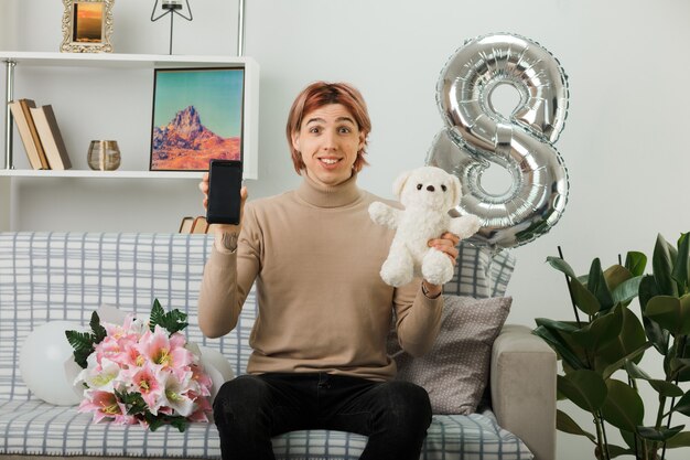 Glimlachende knappe man op gelukkige vrouwendag met teddybeer met telefoon zittend op de bank in de woonkamer