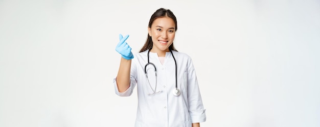 Glimlachende Koreaanse vrouwelijke arts toont een vingerhartgebaar in rubberen handschoenen die een medisch uniform dragen en gelukkig kijken naar de witte achtergrond van de camera