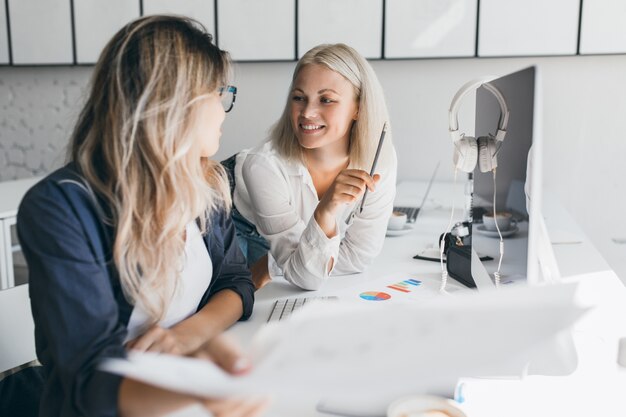 Glimlachende kortharige blonde vrouw praten met collega in kantoor tijdens het spelen met potlood. Binnenportret van vrouwelijke webdesigner die vrolijke vrouw in wit overhemd bekijkt.