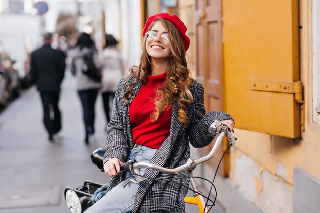 Glimlachende krullende vrouw in rode sweater die op fiets rond stad in koude dag berijdt