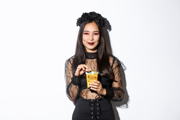 Glimlachende leuke aziatische vrouw die halloween viert, snoepjes houdt en gelukkig grijnst, trick or treat in heksenkostuum.