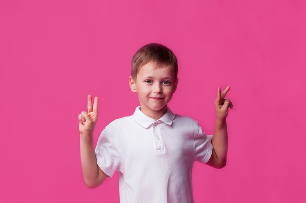 Glimlachende leuke kleine jongen die overwinningsteken op roze achtergrond toont
