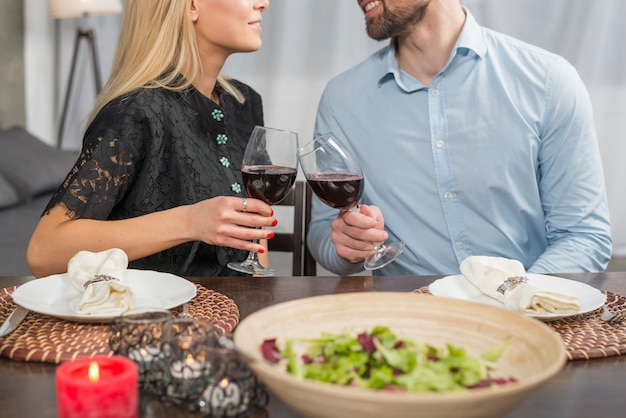 Glimlachende man en vrouw die glazen van drank klinken bij lijst met kom salade en platen