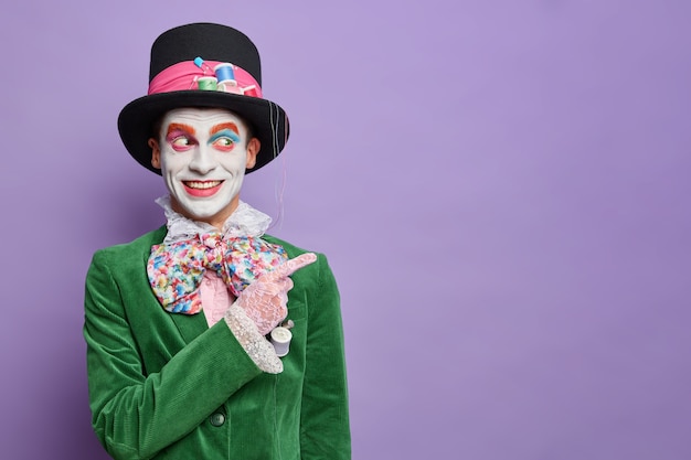 Glimlachende man jurken voor carnaval feest heeft afbeelding van hoedenmaker uit wonderland geeft weg op lege ruimte draagt halloween kostuum en lichte make-up geïsoleerd op paarse muur