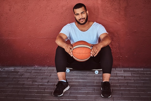 Gratis foto glimlachende man met een donkere huidskleur, gekleed in een wit overhemd en een sportshort, zittend op een skateboard met basketbal en leunend op een muur.