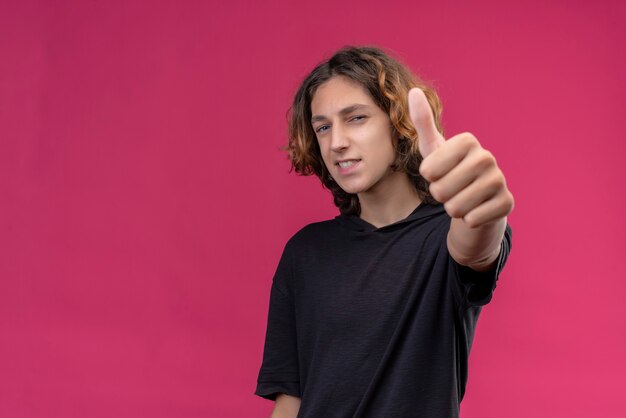 Glimlachende man met lang haar in zwart t-shirt met duim op roze muur