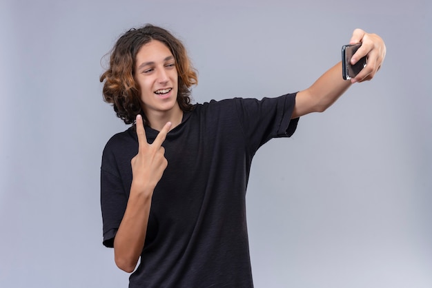 Glimlachende man met lang haar in zwart t-shirt neemt een selfie met gebaar op witte muur