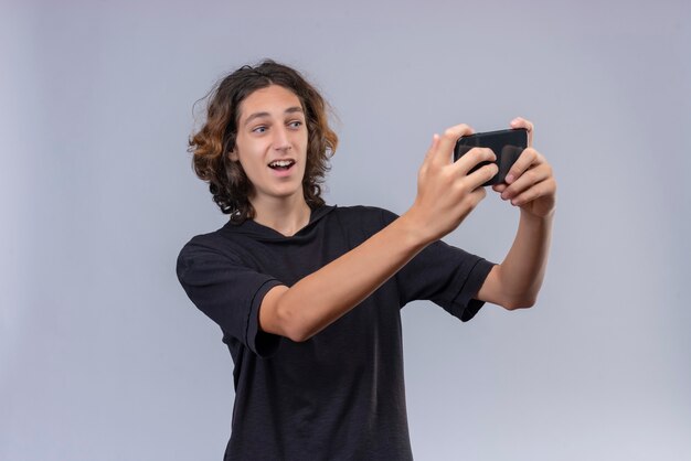 Glimlachende man met lang haar in zwart t-shirt neemt een selfie op witte muur
