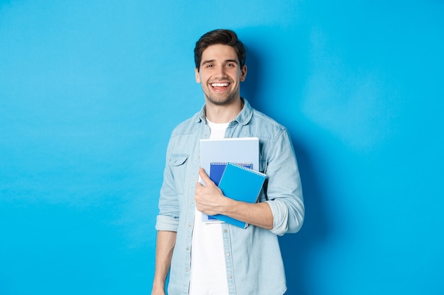 Glimlachende man studeert, houdt notitieboekjes vast en ziet er gelukkig uit, staande over een blauwe achtergrond