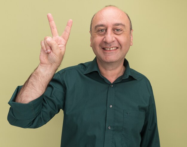 Glimlachende man van middelbare leeftijd die groen t-shirt draagt dat vredesgebaar toont dat op olijfgroene muur wordt geïsoleerd