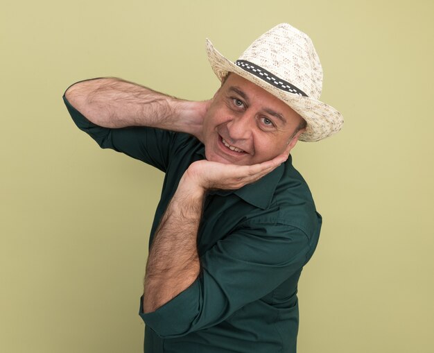 Glimlachende man van middelbare leeftijd met groene t-shirt en hoed greep nek geïsoleerd op olijfgroene muur