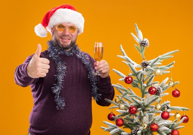 Glimlachende man van middelbare leeftijd met kerstmuts en klatergoud slinger rond de nek met glazen staande in de buurt van versierde kerstboom met glas champagne