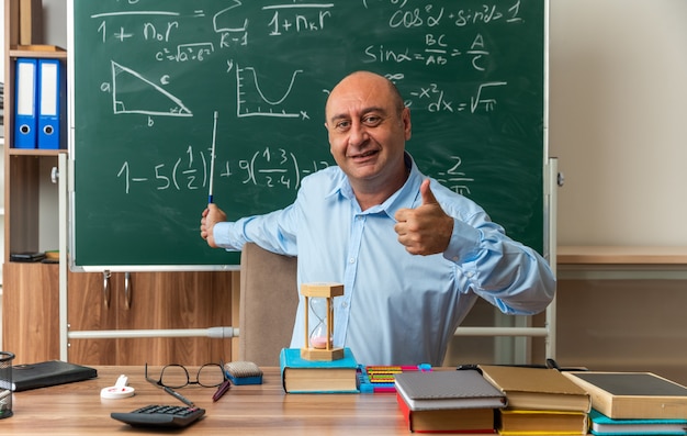 Glimlachende mannelijke leraar van middelbare leeftijd zit aan tafel met schoolhulpmiddelen op het bord met een aanwijzer die de duim in de klas laat zien
