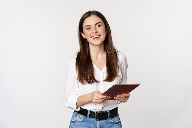 Glimlachende moderne vrouw die met digitale tablet staat, lacht en ziet er gelukkig uit om te poseren tegen ...