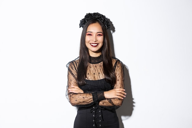 Gratis foto glimlachende mooie aziatische vrouw die halloween viert, die zwarte kroon en kleding, gotische make-up draagt, die gelukkig camera bekijkt. vrouw vieren herfstgebeurtenis, truc of behandelen in kostuum.