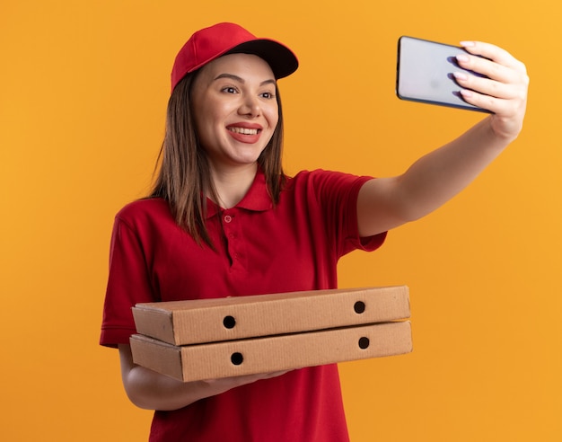 Glimlachende mooie bezorger in uniform houdt pizzadozen vast en kijkt naar telefoon die selfie maakt