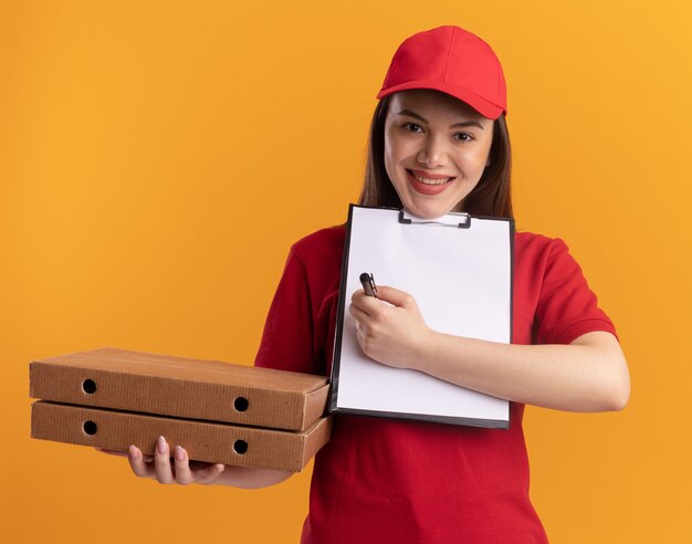 Glimlachende mooie bezorger in uniform houdt pizzadozen vast en schrijft met stift op klembord