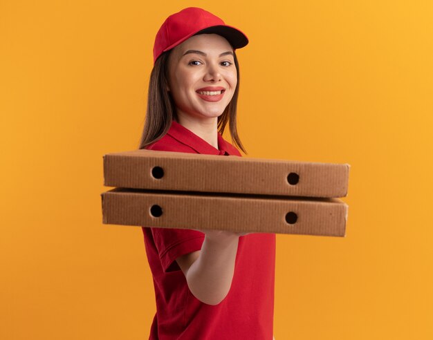 Glimlachende mooie bezorger in uniform staat zijwaarts pizzadozen op oranje te houden