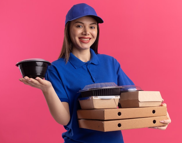 Glimlachende mooie bezorgvrouw in eenvormig voedselpakket en containers op pizzadozen die op roze muur met exemplaarruimte worden geïsoleerd