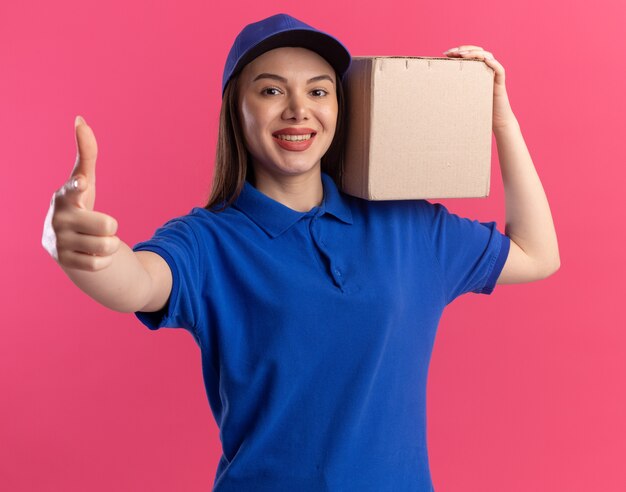 Glimlachende mooie bezorgvrouw in uniform beduimelt omhoog en houdt kartonnen doos op de schouder op roze