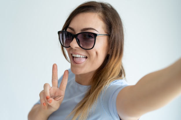 Glimlachende mooie vrouw die selfie foto neemt, overwinningsteken toont en camera bekijkt.