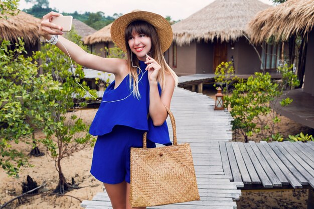 Glimlachende mooie vrouw die zelfportret maakt via de mobiele telefoon op haar tropische vakantie in Thailand. Zomer felblauwe kleding ^ trendy strohoed en tas. Rode lippen. Blije stemming.