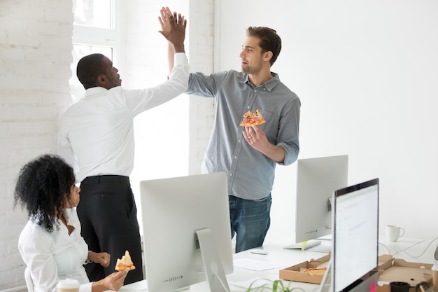 Glimlachende multiraciale medewerkers die high-five geven pizza samen eten op kantoor