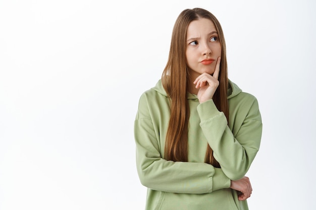 Gratis foto glimlachende nadenkende vrouw denkend, opzij kijkend naar logo met peinzend gezicht, staande tegen een witte muur, nadenkend