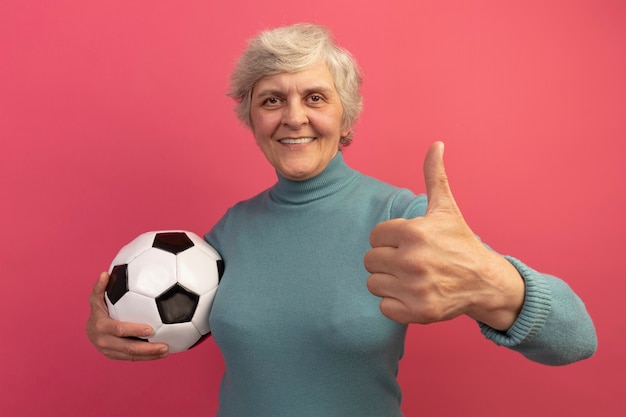 Glimlachende oude vrouw die een blauwe coltrui draagt en voetbal vasthoudt en naar de voorkant kijkt met duim omhoog geïsoleerd op roze muur
