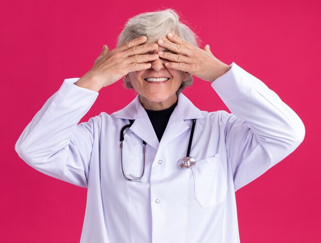 Glimlachende oudere vrouw in doktersuniform met stethoscoop die ogen bedekt met handen