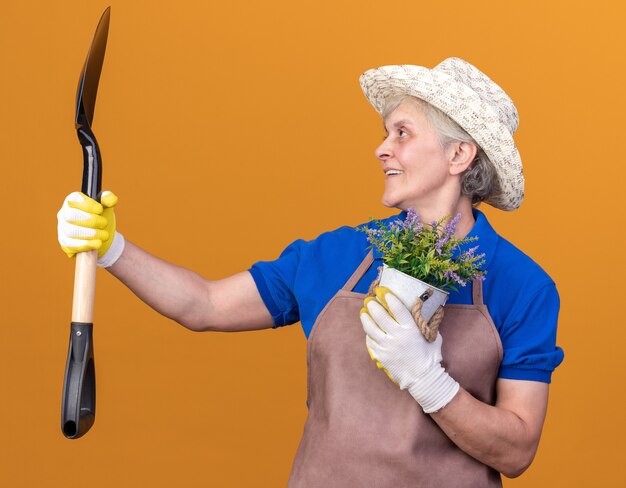 Glimlachende oudere vrouwelijke tuinman met een tuinhoed en handschoenen die een bloempot vasthoudt en naar een schop kijkt