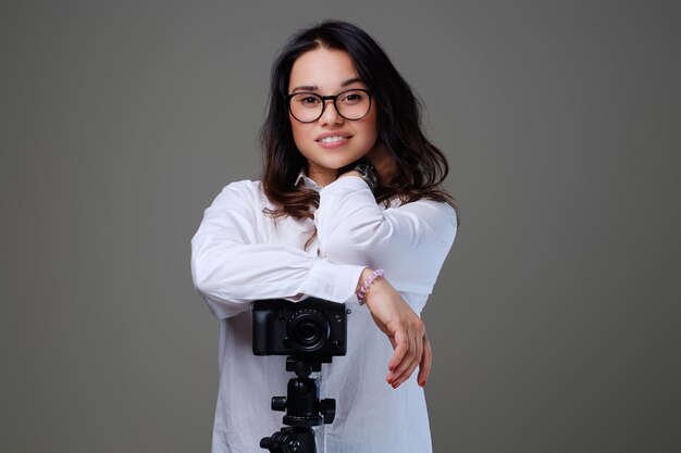 Glimlachende, positieve brunette vrouw in brillen die foto's maken met een professionele fotocamera. Geïsoleerd op grijze achtergrond.