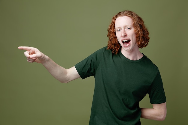 Glimlachende punten aan de zijkant jonge knappe kerel met een groen t-shirt geïsoleerd op een groene achtergrond