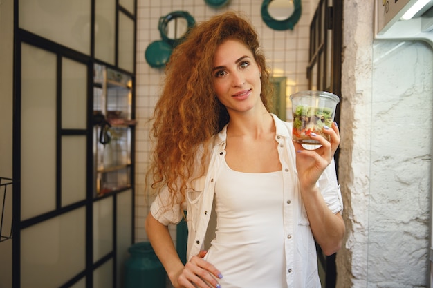 Glimlachende roodharige jonge dame die zich in koffie bevindt terwijl het houden van salade.