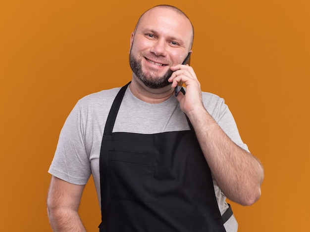 Glimlachende slavische mannelijke kapper van middelbare leeftijd in uniform spreekt over telefoon die op oranje muur wordt geïsoleerd