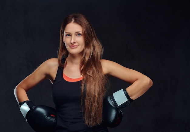 Glimlachende sportieve vrouw gekleed in sportkleding dragen van bokshandschoenen poseren in een studio. Geïsoleerd op een donkere gestructureerde achtergrond.