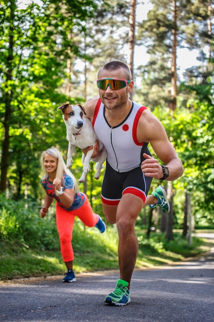 Glimlachende sportman in zonnebril op de vlucht met kleine hond in zijn hand. Lopende vrouw op de achtergrond.