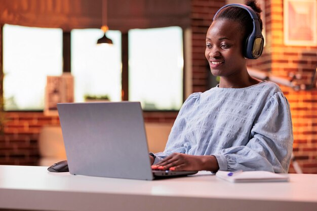 Glimlachende student in hoofdtelefoons die online lessen op laptop thuis bijwoont. Afro-Amerikaanse vrouw kijkt naar educatieve video in moderne kamer met grote ramen en prachtig warm zonsonderganglicht