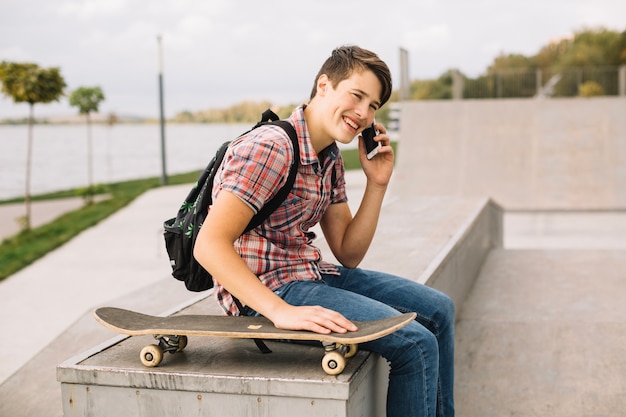 Gratis foto glimlachende tiener die op telefoon dichtbij skateboard spreekt