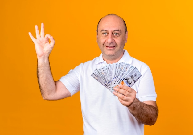 Glimlachende toevallige volwassen man die contant geld houdt en ok gebaar toont dat op gele muur wordt geïsoleerd
