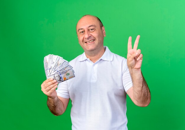Glimlachende toevallige volwassen mens die contant geld houdt en vredesgebaar toont dat op groene muur wordt geïsoleerd